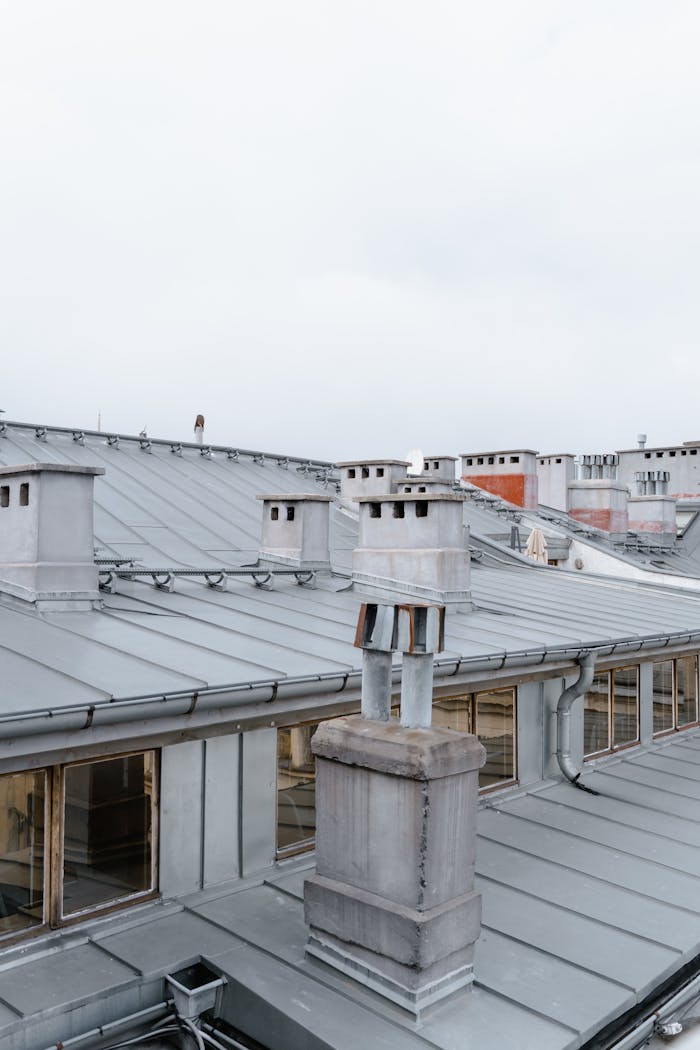 Detailed view of city rooftops with metal chimneys and windows, under cloudy sky.